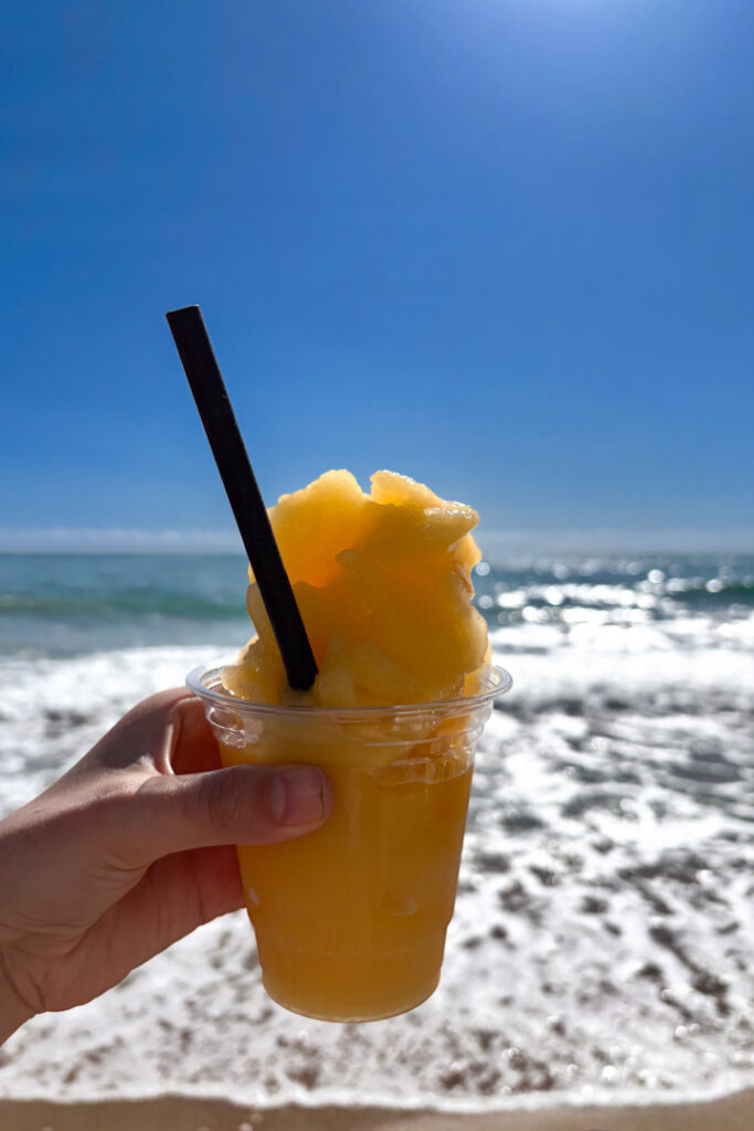 Eine Hand, die einen gelben Frozen Daiquiri vor dem Meer hält