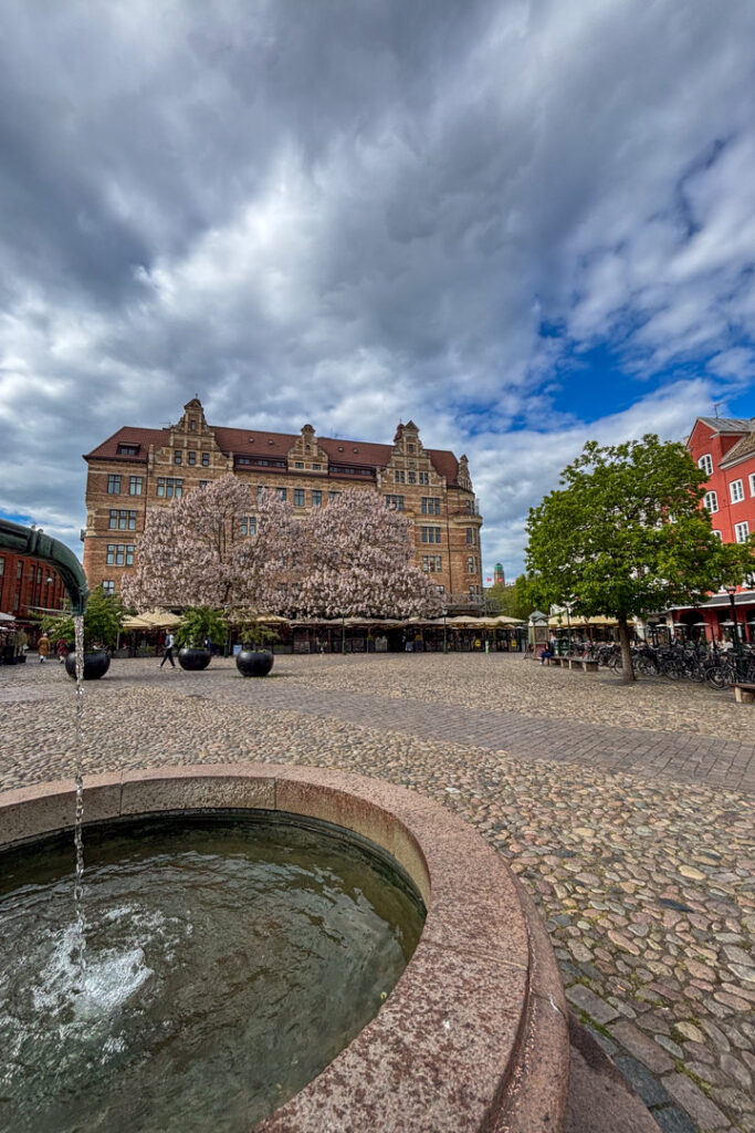 Ein Platz mit einem Brunnen, Bäumen und einem Gebäude