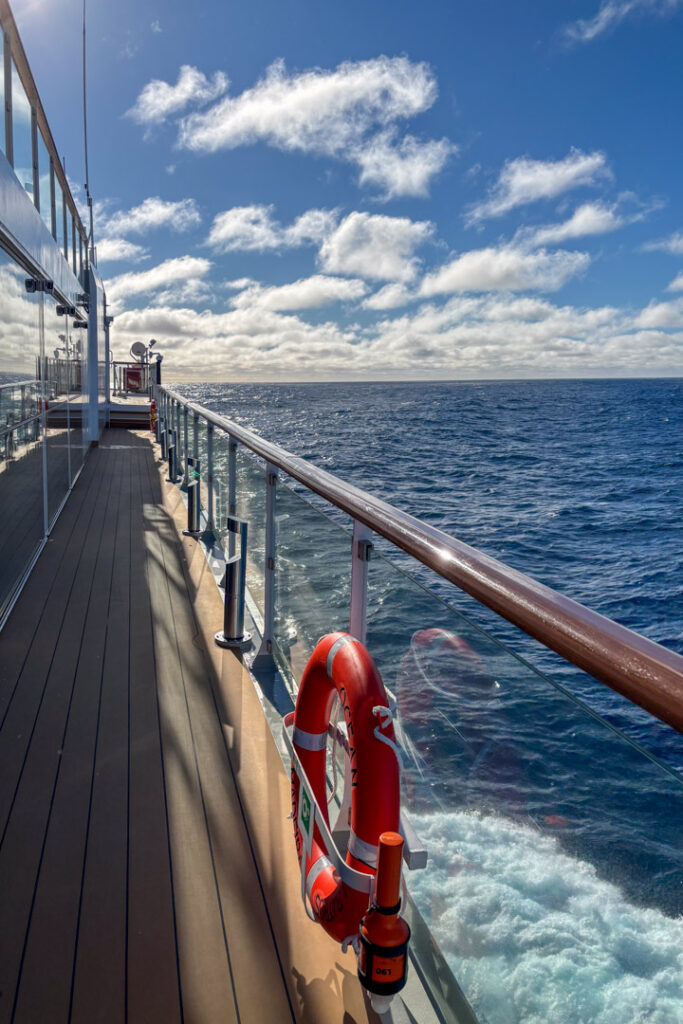 Blick entlang eines hölzernen Schiffsdecks bei Sonnenschein auf das offene, tiefblaue Meer unter einem leicht bewölkten Himmel, mit einem roten Rettungsring an der Reling im Vordergrund