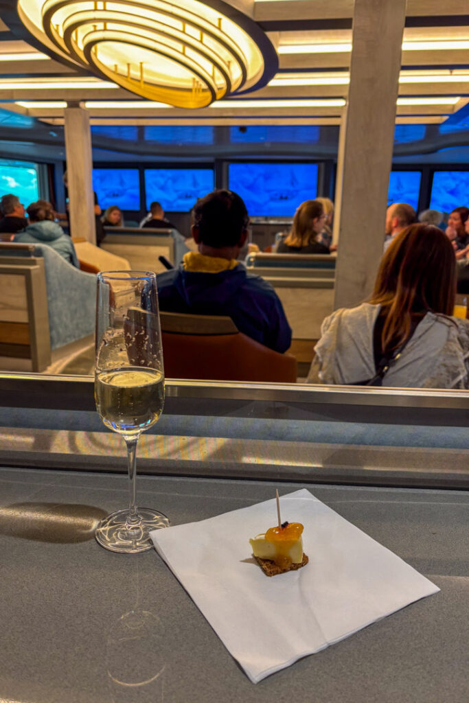 Ein Glas Sekt und Häppchen in einer Lounge, im Hintergrund hält jemand eine Rede