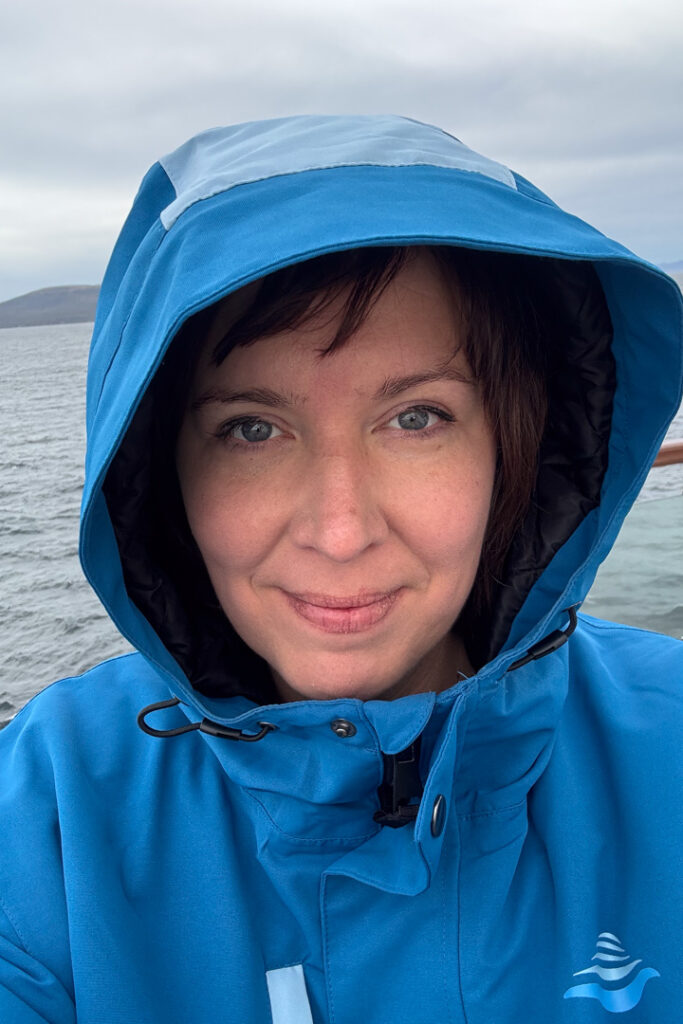 Eine Frau strahlt bei einem Selfie in die Kamera, sie hat eine blaue Jacke mit Kapuze an