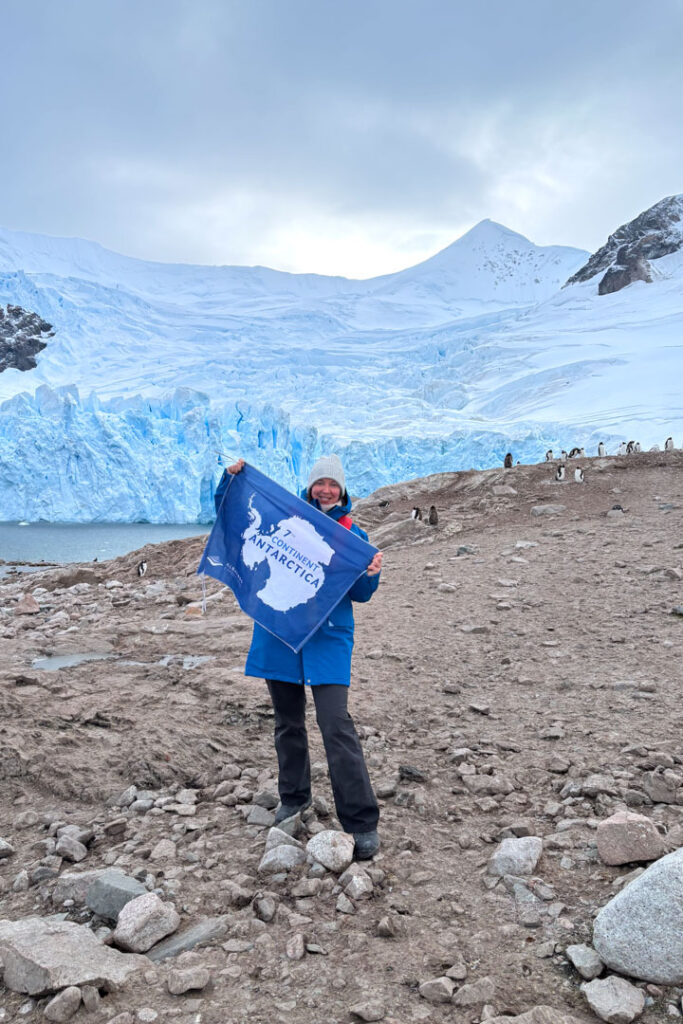 Eine Frau mit dicken Winterklamotten steht mit einer Flagge von der Antarktis "7th Continent" vor einem Gletscher und Pinguinkolonie