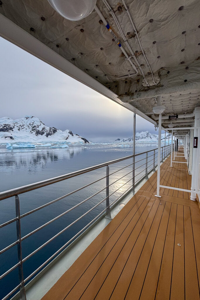 Blick von einem überdachten Schiffsdeck mit Holzplanken auf eine spiegelglatte, antarktische Meeresbucht, in der sich verschneite Berge und hellblaue Eisberge unter einem bewölkten Himmel spiegeln