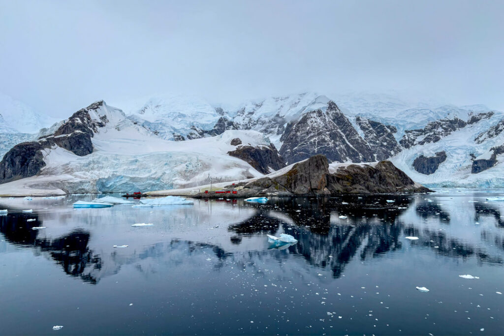 Eine weitläufige arktische oder antarktische Küstenlandschaft unter einem wolkenverhangenen, blassgrauen Himmel. Im Hintergrund erheben sich mächtige, schroffe Berge und Gletscher, die von tiefen Schneefeldern und bläulichem Eis bedeckt sind, wobei dunkle Felswände durchbrechen