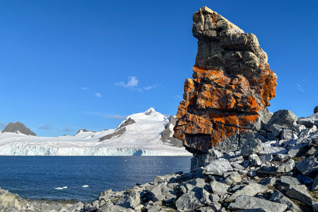 Eine markante, aufragende Felsformation, die dicht mit leuchtend orangeroter Flechte bewachsen ist, steht im Vordergrund einer steinigen antarktischen Küste vor tiefblauem Meer, einer gewaltigen Gletscherfront und schneebedeckten Bergen unter strahlend blauem Himmel