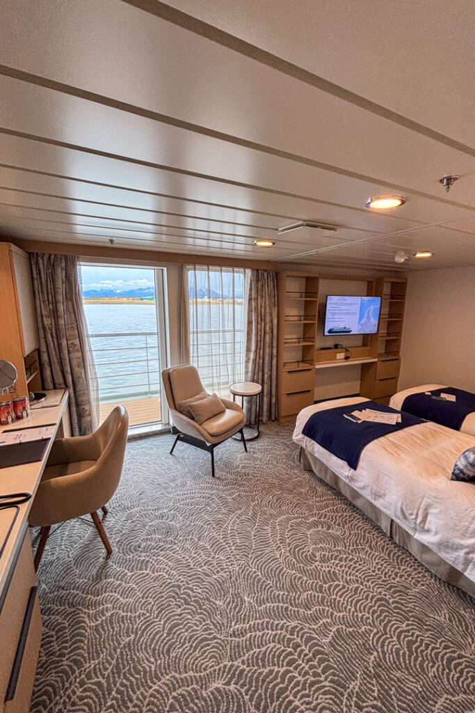 Eine große Kabine auf einem Schiff mit hellem Teppich, 2 Einzelbetten, einem TV, einem Schreibtisch mit Stuhl und großem Balkon