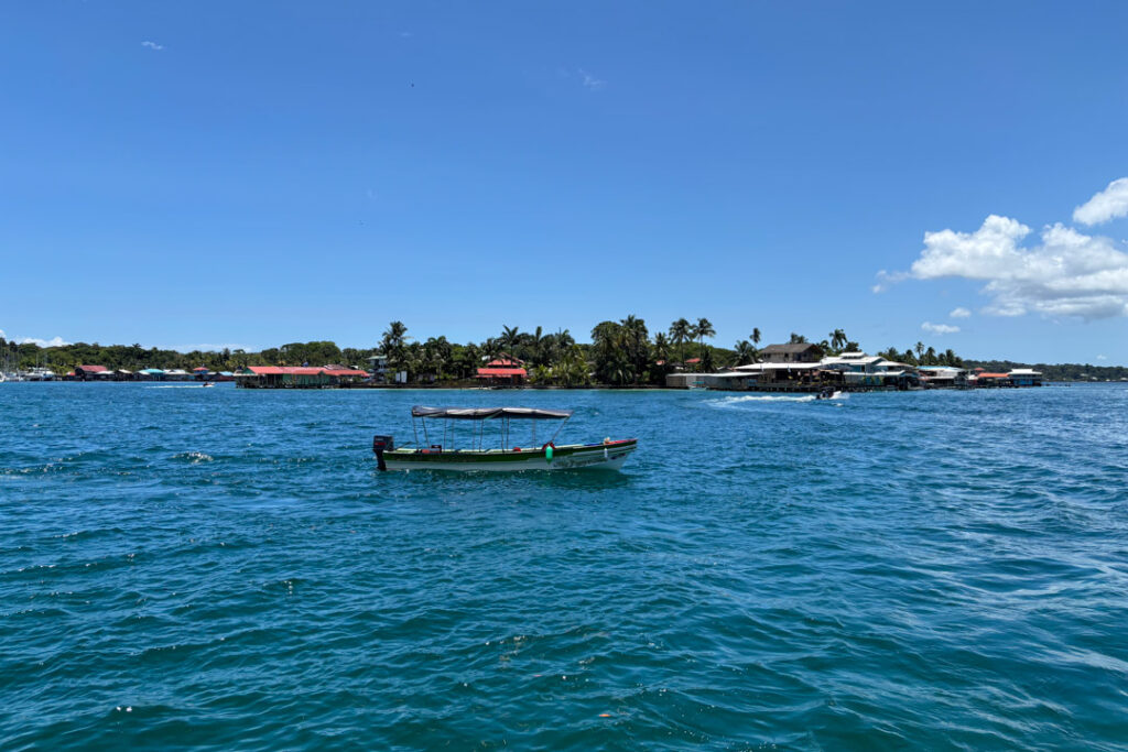 Ein kleines, überdachtes Wassertaxi mit grünem Rand liegt mitten auf dem tiefblauen Meer vor Bocas Town. Im Hintergrund erstreckt sich die flache Küstenlinie der Isla Colón mit bunten Holzhäusern, Palmen und Bootsstegen unter einem strahlend blauen Himmel mit vereinzelten weißen Wolken