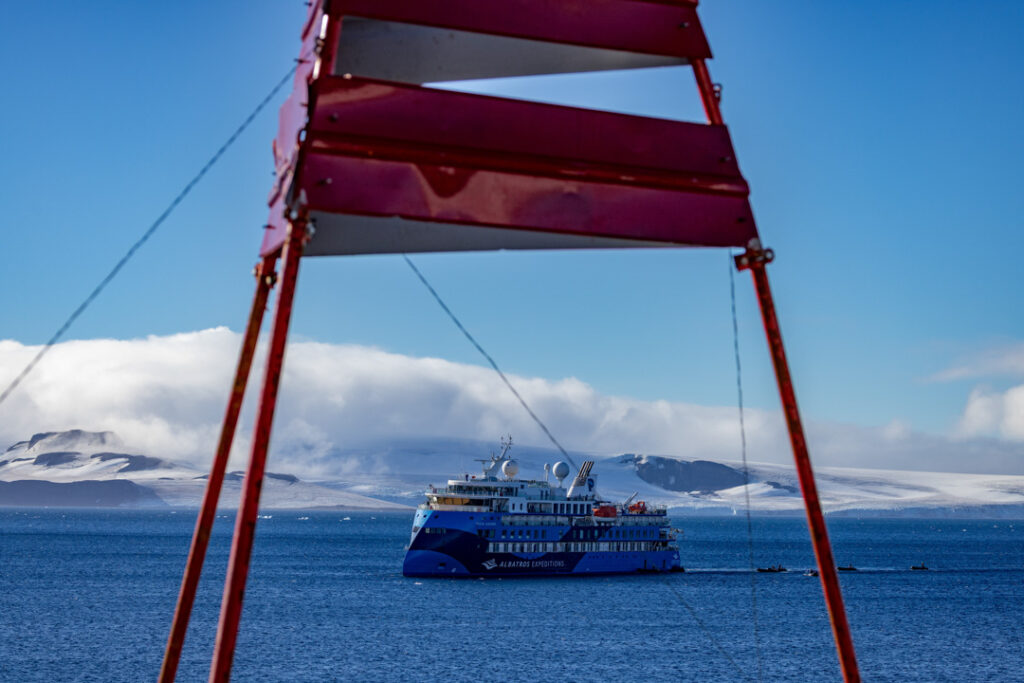 Ein blau-weißes Expeditionsschiff mit der Aufschrift „Albatros Expeditions“ fährt auf dem Meer vor einer schneebedeckten Küste, fotografiert durch die Beine eines roten Metallgerüsts im Vordergrund