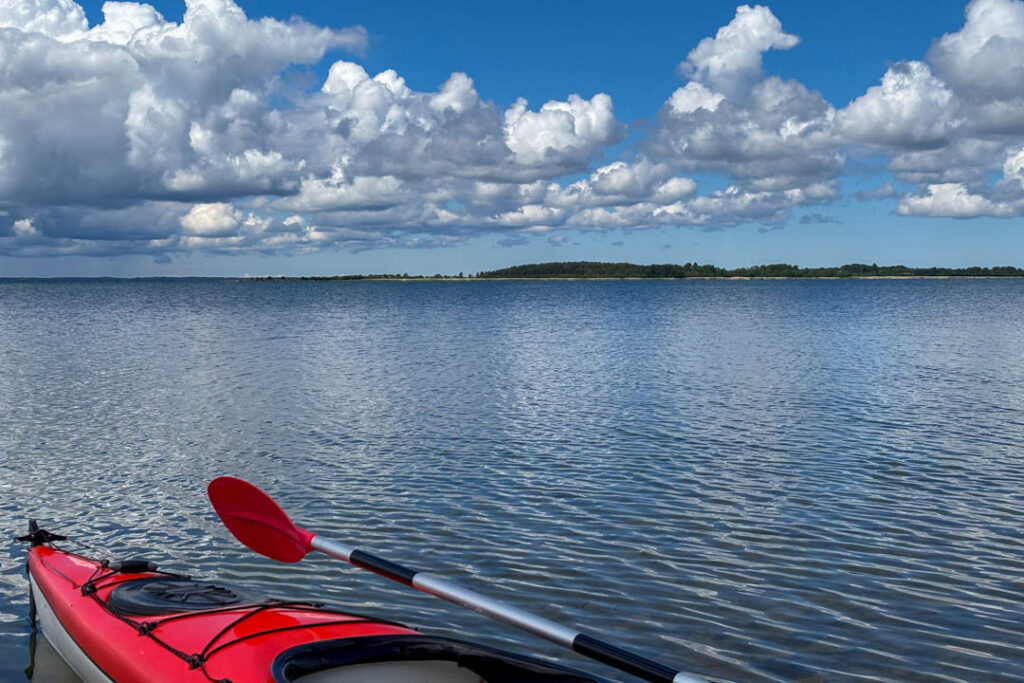 Blick aus der Perspektive eines roten Kajaks mit Doppelpaddel auf eine ruhige, weite Wasserfläche unter einem leicht bewölkten blauen Himmel mit einer bewaldeten Insel im Hintergrund