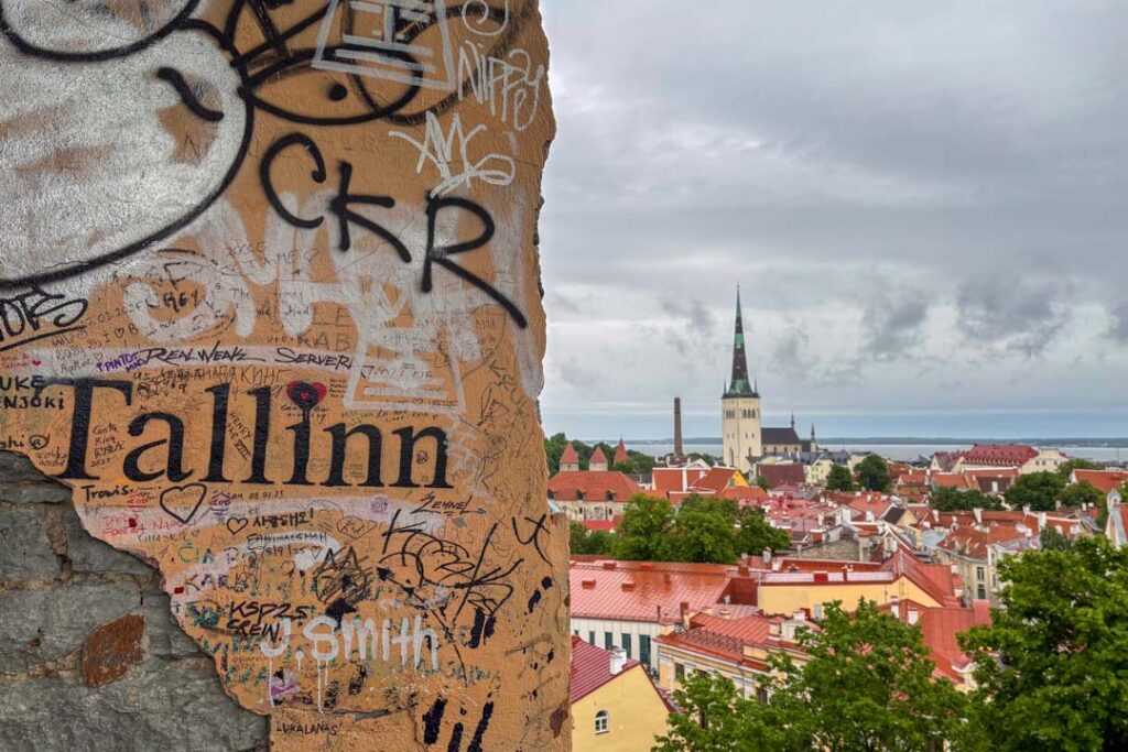 Blick auf eine Mauer mit Graffiti und dem Namen "Tallinn" und dahinter die Altstadt und im Hintergrund die Ostsee