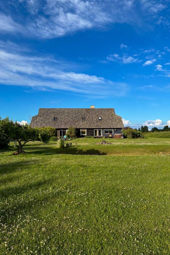 Ein langes, traditionelles Holzhaus mit einem großen Schilf- oder Reetdach auf einer weitläufigen, grünen Wiese unter blauem Himmel