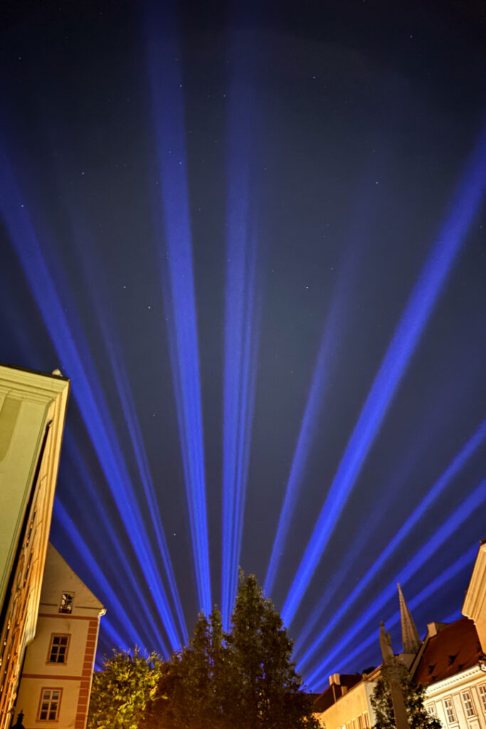 Zahlreiche intensive, blaue Lichtstrahlen ragen fächerförmig zwischen historischen Hauswänden empor in den nächtlichen Sternenhimmel