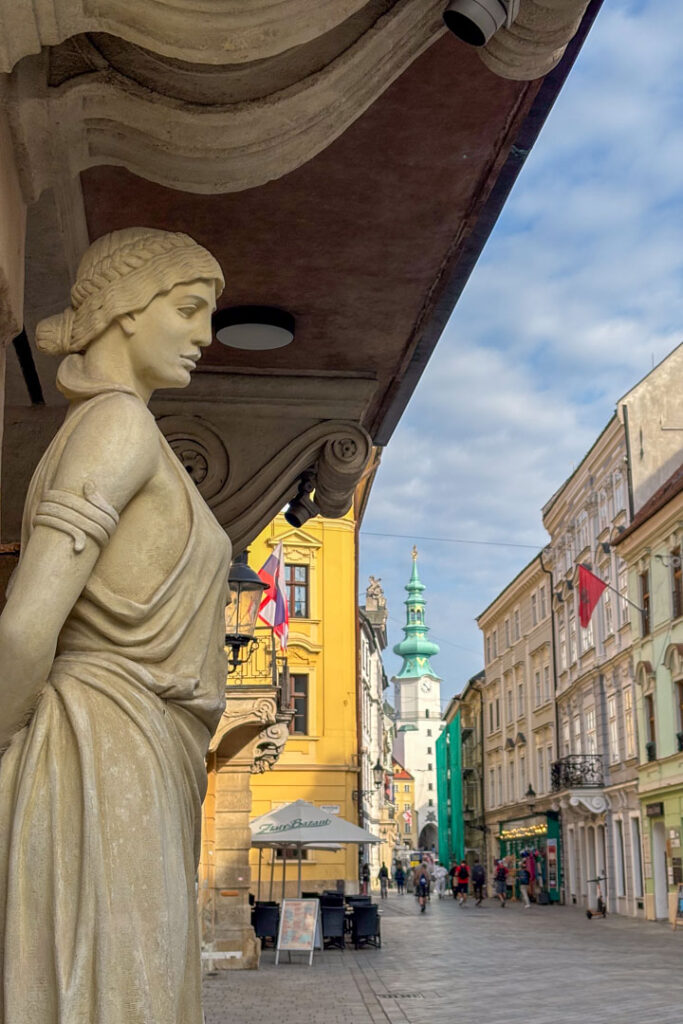 Nahaufnahme einer steinernen Frauenstatue an einer Hausfassade im Vordergrund, mit Blick auf eine belebte Einkaufsstraße in Bratislava und das Michaelertor im Hintergrund