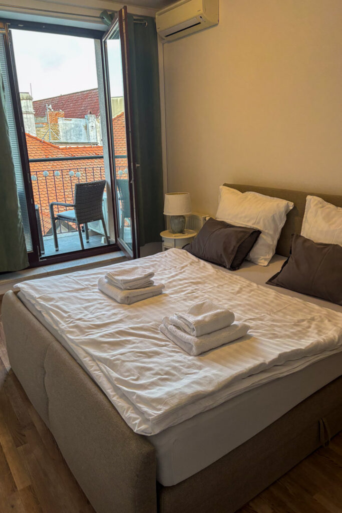 Ein einladendes, frisch bezogenes Doppelbett mit weißen Handtüchern in einem Hotelzimmer, im Hintergrund öffnet sich eine Balkontür mit Blick auf rote Ziegeldächer