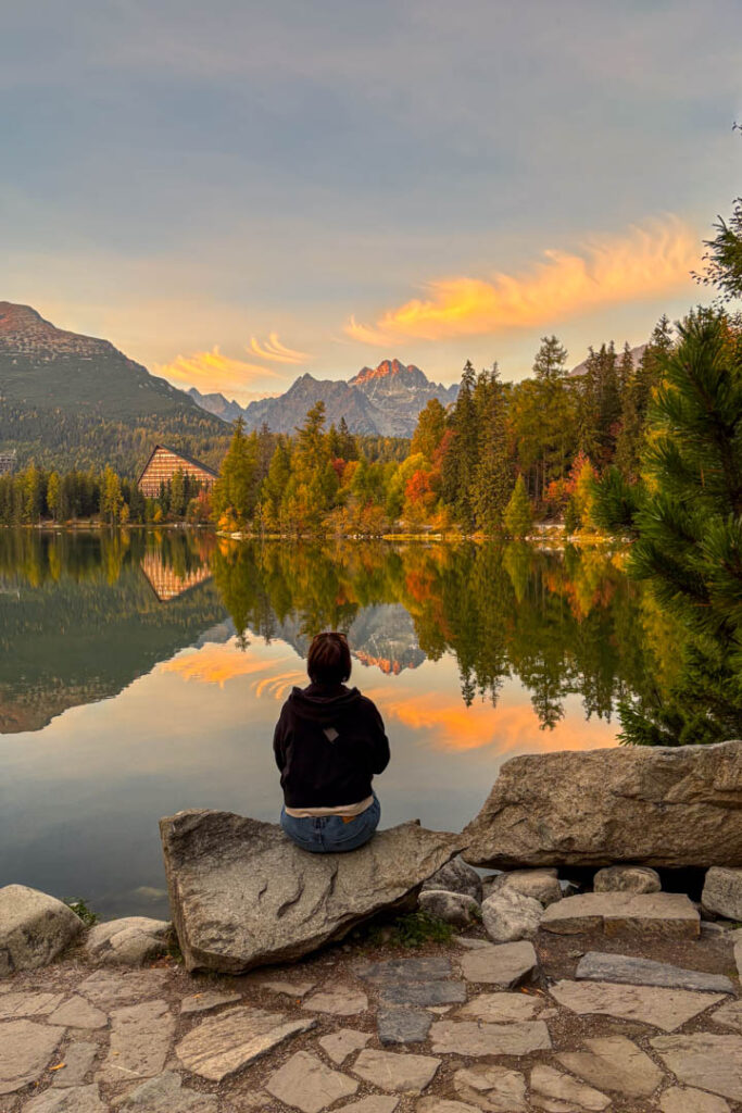 Eine Person sitzt von hinten fotografiert auf einem Felsen am Ufer des Bergsees Štrbské Pleso und blickt auf die herbstlichen Bäume und die von der Abendsonne erleuchteten Gipfel der Hohen Tatra