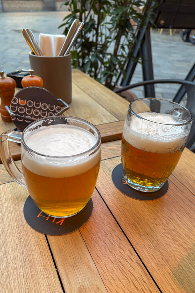 Zwei Gläser frisch gezapftes, helles Bier stehen auf Bierdeckeln auf einem hölzernen Tisch im Außenbereich eines Restaurants