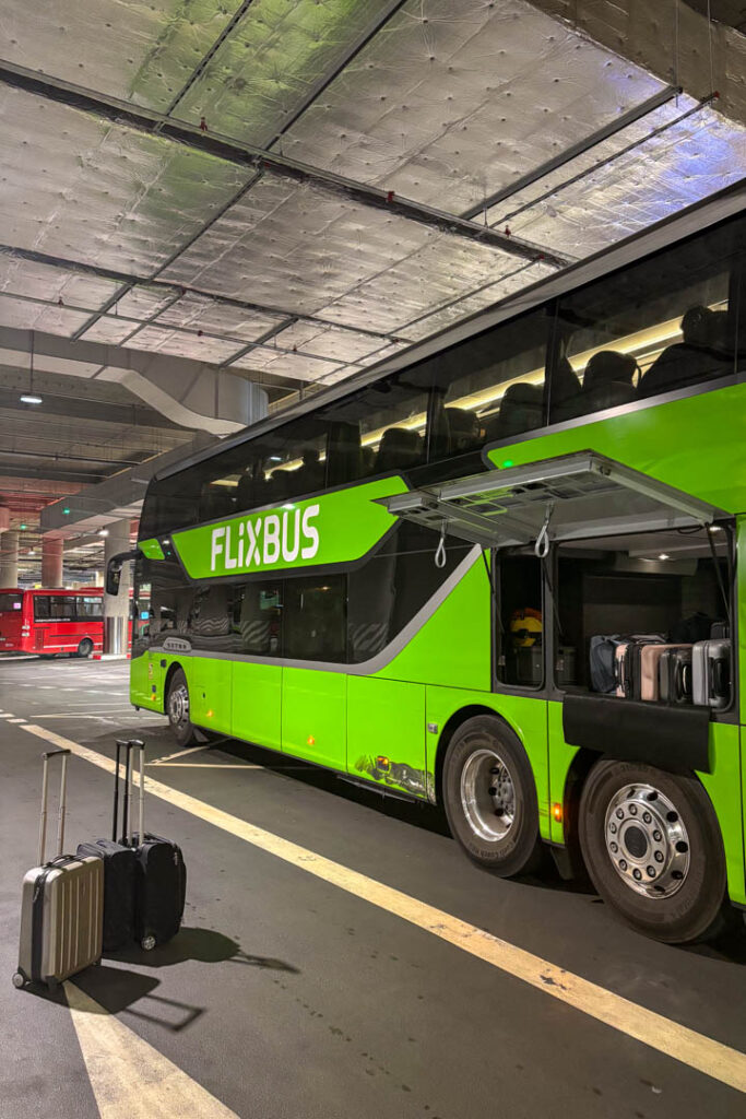 Ein grüner FlixBus-Doppeldeckerbus parkt mit geöffneter Gepäckklappe in einem Busbahnhof, daneben stehen zwei Rollkoffer auf dem Asphalt bereit