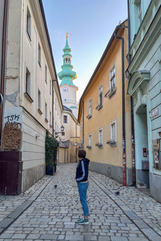 • Eine Person steht in einer gepflasterten Gasse in Bratislava und blickt hinauf zum markanten grünen Turm des Michaelertors