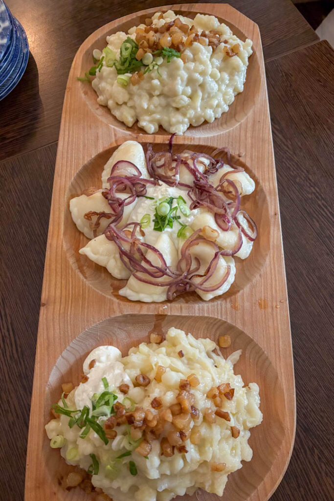 Eine hölzerne Servierplatte mit drei Mulden, gefüllt mit traditionellen slowakischen Gerichten wie Bryndzové Halušky und Piroggen, garniert mit Speck und Frühlingszwiebeln