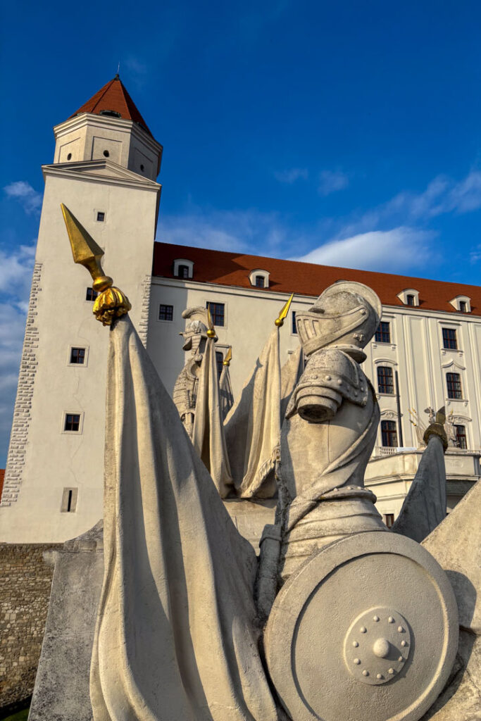 Nahaufnahme einer historischen Ritterstatue mit Schild vor dem weißen Turm und der Fassade der Burg Bratislava unter blauem Himmel