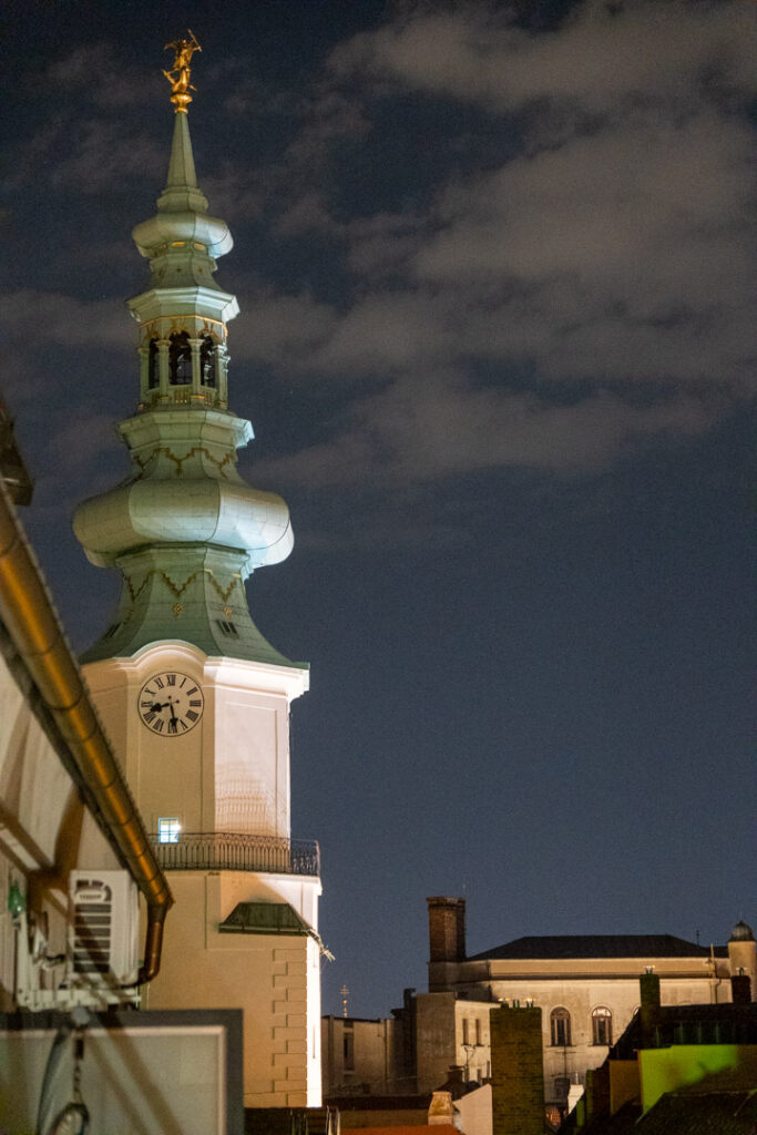 Der beleuchtete barocke Turm des Michaelertors bei Nacht vor einem leicht bewölkten Himmel in Bratislava