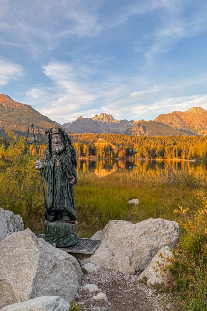 Eine geschnitzte Holzfigur am Ufer des Bergsees Štrbské Pleso in der Hohen Tatra, im Hintergrund spiegelt sich das markante Hotelgebäude vor den Bergen