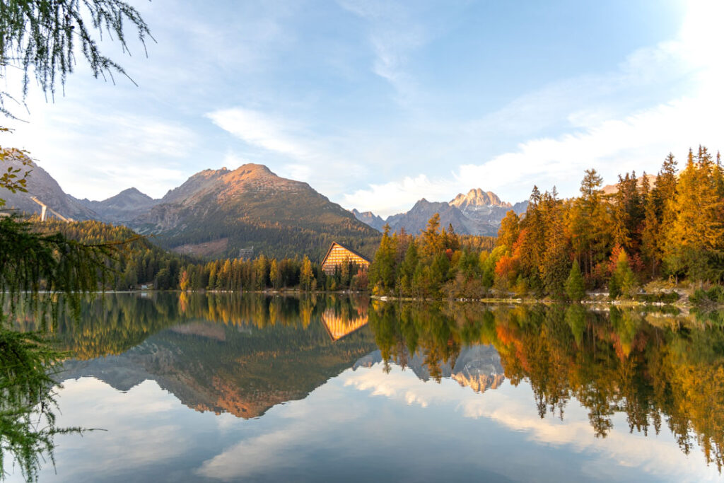 Landschaftsaufnahme des Bergsees Štrbské Pleso in der Hohen Tatra, gerahmt von Nadelbäumen, in dessen klarem Wasser sich die herbstlichen Wälder und Berge spiegeln